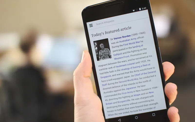 A hand holding a Nexus 6 with the Wikipedia mobile homepage displayed, in an office setting