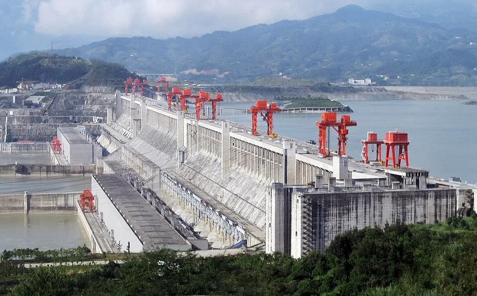 A huge concrete dam with red cranes along its top, holding back a wide river between green hills.