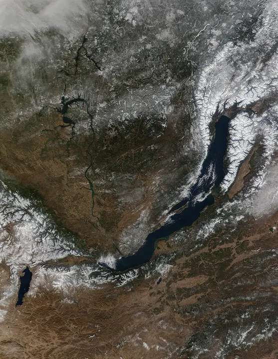 A satellite view of a long, narrow dark-blue lake lying among snow-streaked brown mountains.