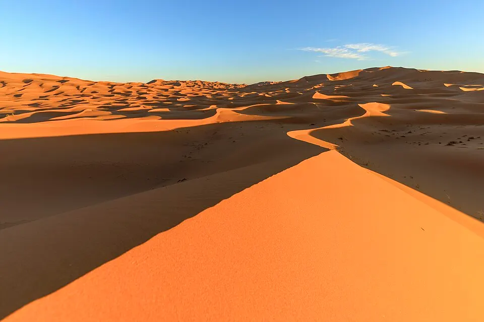 Smooth orange sand dunes with sharp curving ridges under a clear blue sky at sunset.