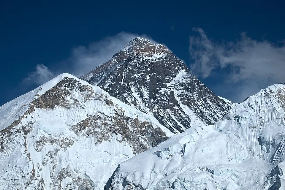 The snow-streaked dark rocky peak of a very high mountain against a deep blue sky, with snowy ridges in front.