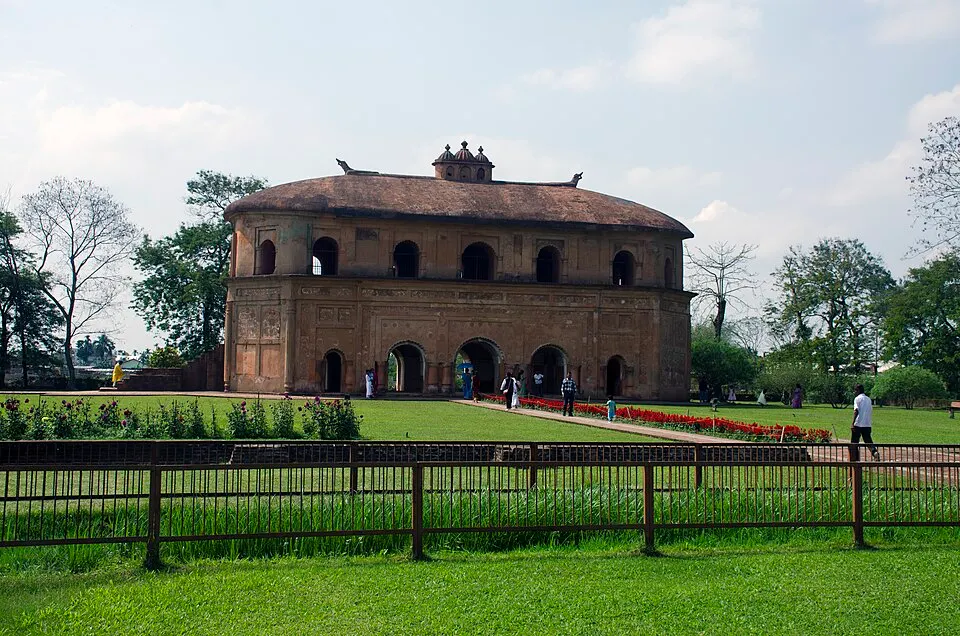 A two-storey brick pavilion with arched openings and a curved roof topped by small ornaments stands in a green garden.
