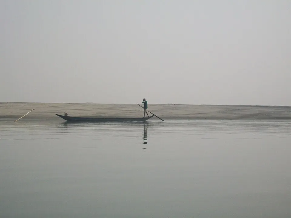 A man poles a long narrow wooden boat across a calm, misty river beside a flat sandy bank.