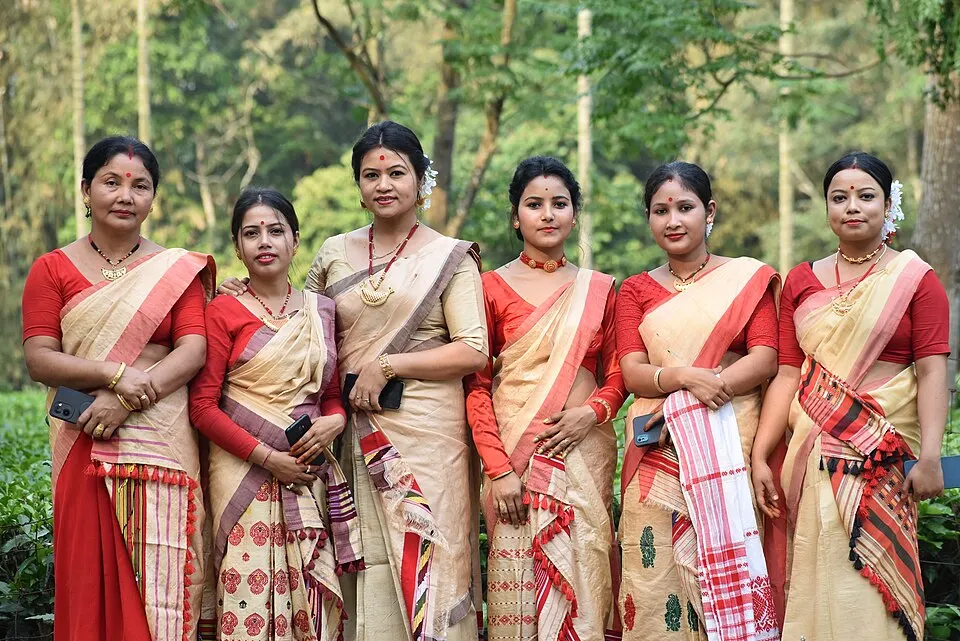 Six women in cream-and-red handwoven mekhela chador, with flowers in their hair, stand together in front of green trees.