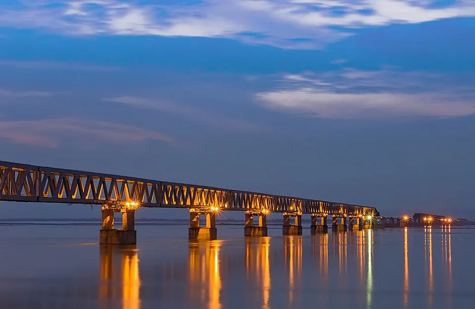 A long steel truss bridge on stone pillars, lit with warm lights, crosses a very wide river at dusk.