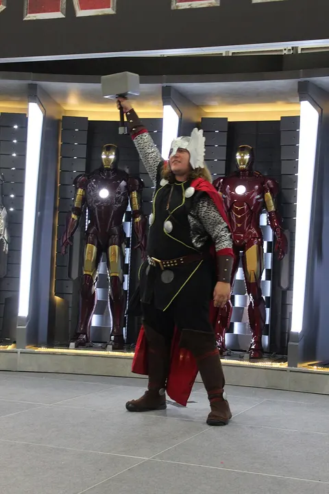 A cosplayer in a winged silver helmet and red cape raising a large hammer overhead, with two red and gold armour suits behind.