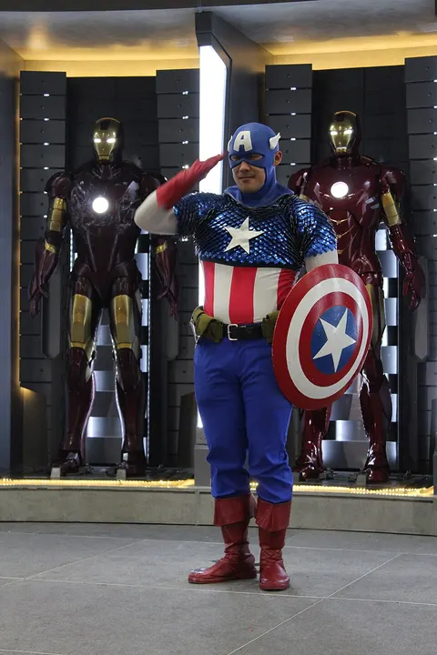 A cosplayer in a blue costume with a star on the chest and a winged cowl, holding a round red, white and blue shield and saluting.