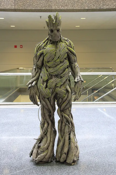 A cosplayer in a tall costume made of grey-brown tree bark and moss, with a stern wooden face, standing in a hall.
