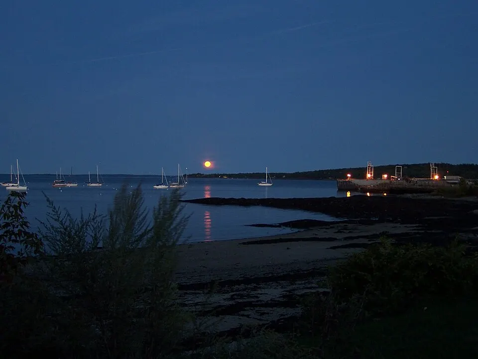 A large orange full moon rising low over a calm harbour with sailboats at dusk.