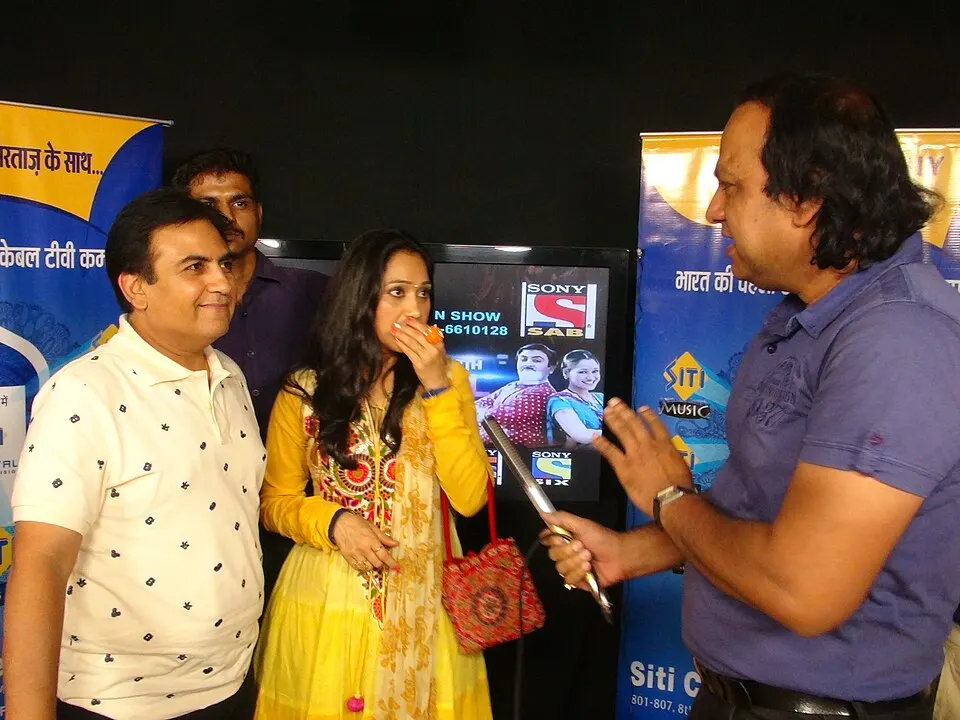 A man in a white polo and a woman in a yellow kurta are interviewed by a man holding a microphone in front of event banners.