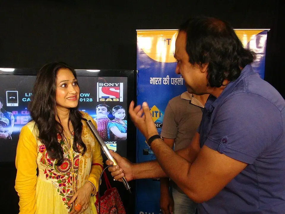 A smiling woman in a yellow embroidered kurta is interviewed by a man with a microphone in front of a TV screen and a banner.