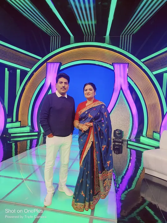 A man in a navy sweater and white trousers stands beside a woman in a blue saree on a bright neon-lit studio set.