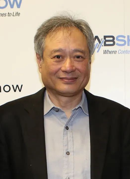 A smiling grey-haired man in a dark jacket and pale blue shirt stands in front of an event backdrop.