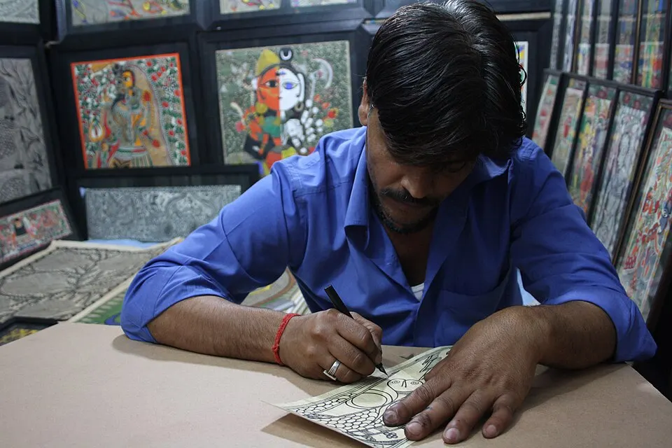 एक कलाकार काग़ज़ पर कलम से रंग-बिरंगी लोक चित्रकला की रेखाएँ बनाता हुआ, पीछे कई चित्र टँगे हैं।