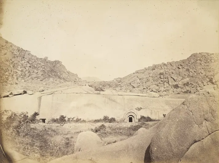 An old sepia photograph shows a long smooth rock face with a small arched doorway, among rocky boulder-strewn hills.