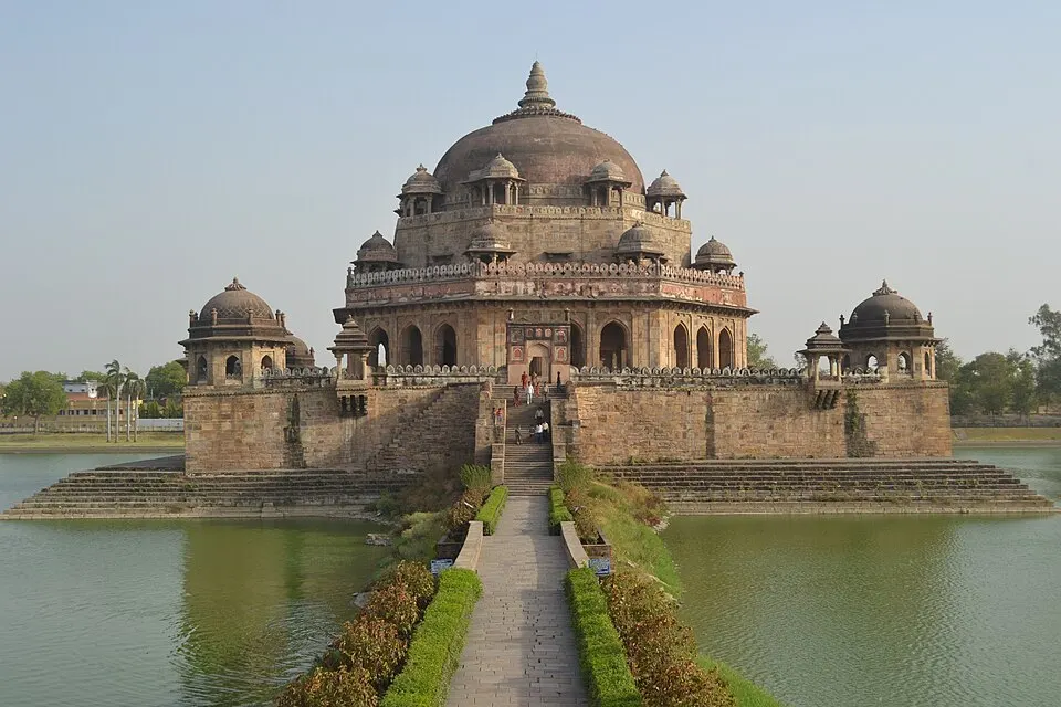 A large domed sandstone tomb with small corner pavilions stands on a platform in the middle of a lake, reached by a paved causeway.