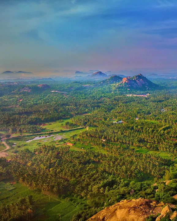Green coconut groves and farmland seen from a rocky hilltop, with pinkish granite boulders on the hills in the distance.