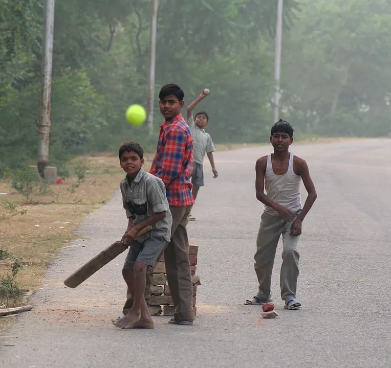 Four boys play cricket on an empty village road, one batting with a plank bat in front of a brick-stack wicket while a yellow ball flies past.