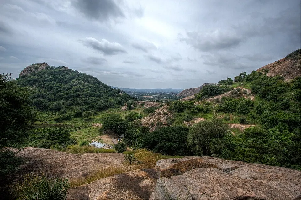 Boulder-strewn green hills and scrub under a stormy sky, seen from a rocky outcrop.