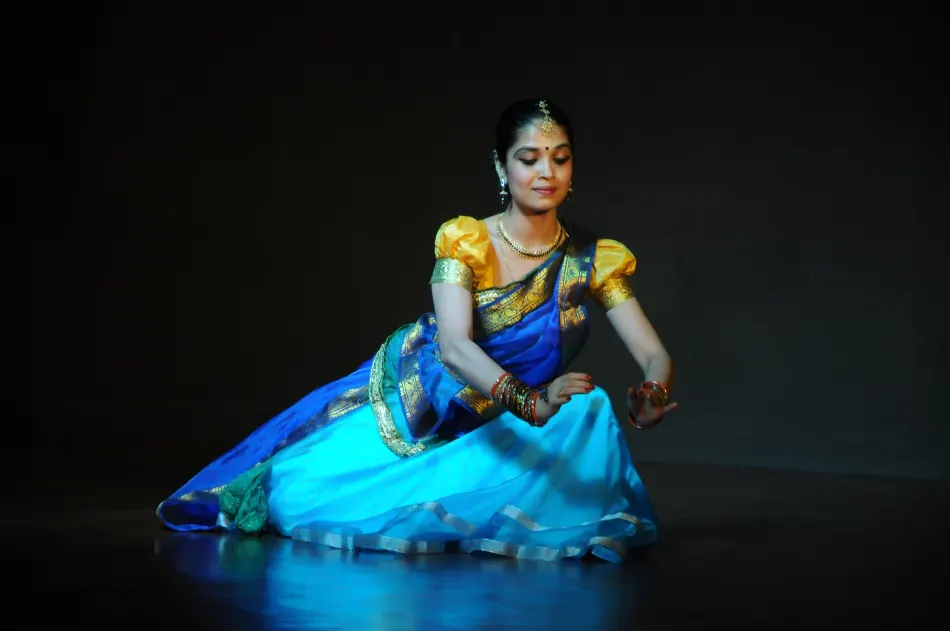 A classical dancer in a blue and yellow costume with gold jewellery poses on a dark stage.