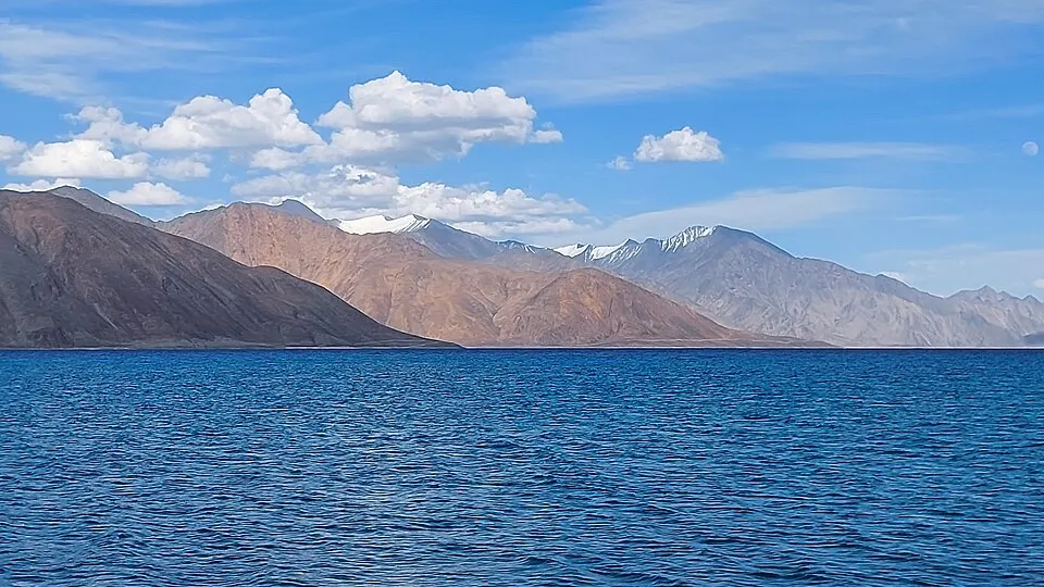 A deep blue mountain lake with brown and snow-capped peaks beyond under a bright sky with clouds.