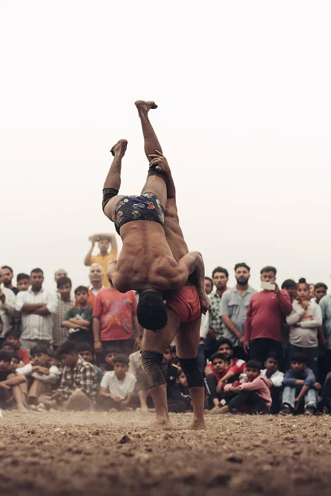 Two wrestlers in an earthen ring, one lifted upside-down over the other's shoulder, as a crowd watches.