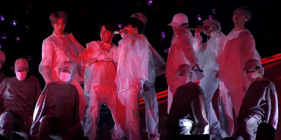 Performers in white outfits sing on stage with masked dancers crouching in the foreground under red light.