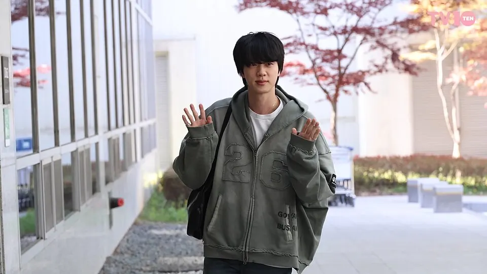 A young man with dark hair in a grey-green hoodie waves both hands while walking outdoors.
