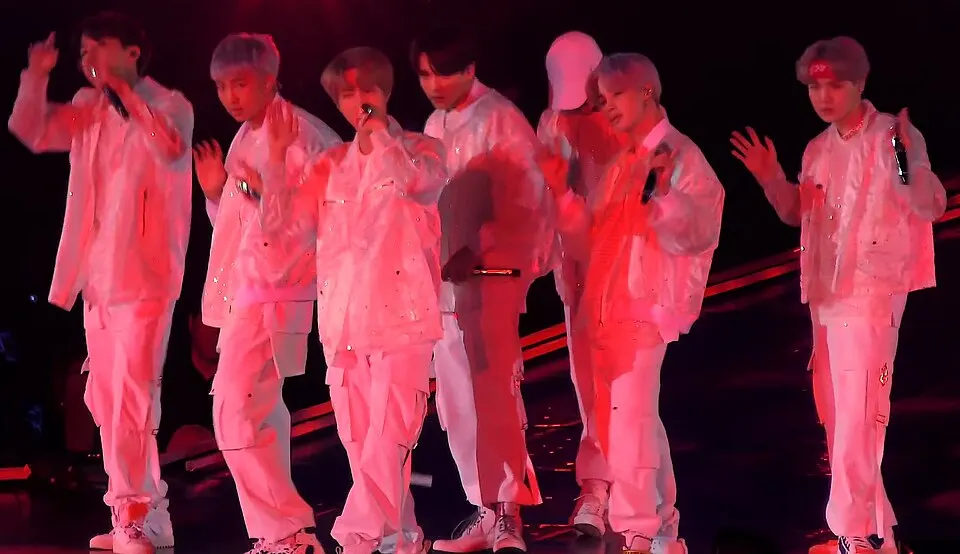 Seven performers in white and pink outfits dance on a dark stage lit in red.