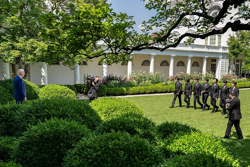 A man in a blue suit watches as a group of men in black suits walk across a lawn beside a white building while photographers film.