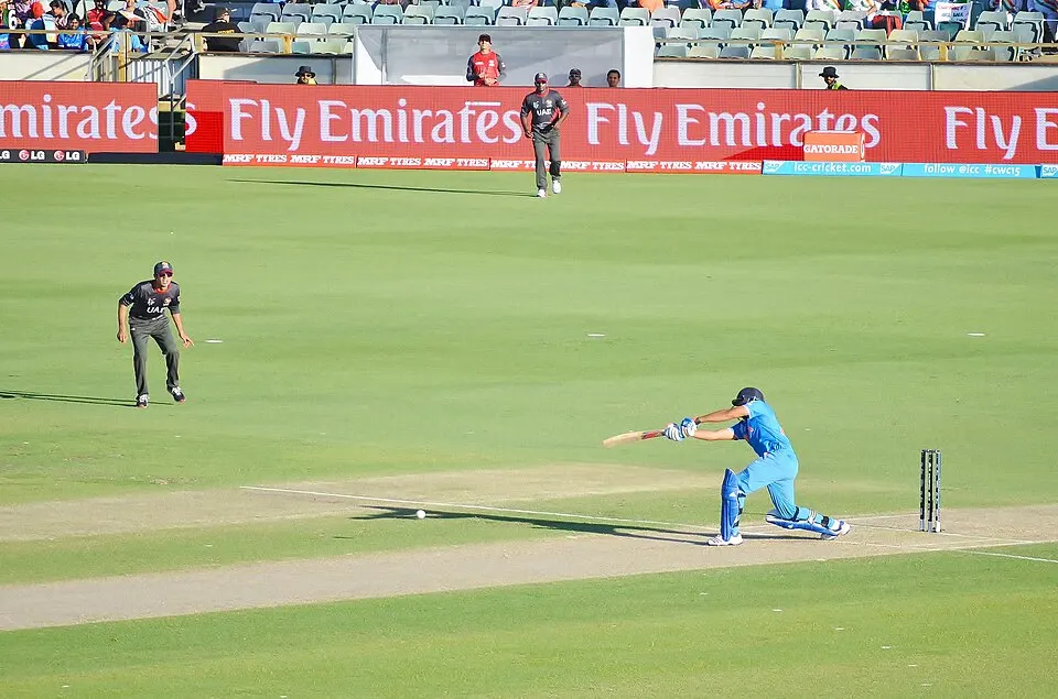 A batsman in blue kit plays a shot while fielders in dark shirts stand around him on a green field.