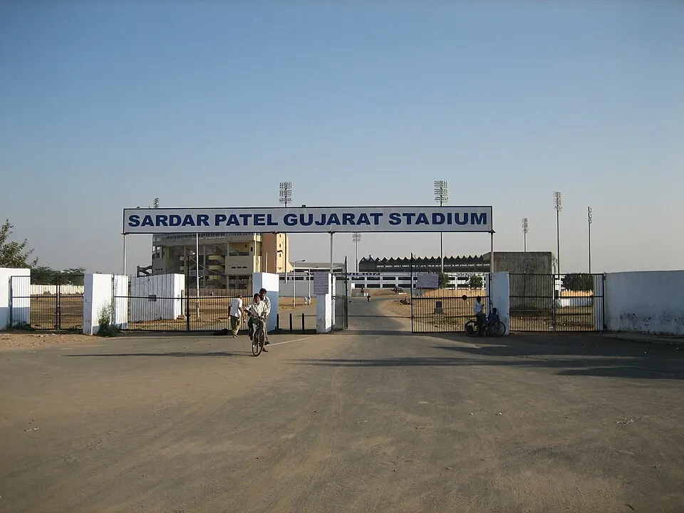 A large gate with a blue sign above a dusty road, with a stadium and floodlight towers behind it.