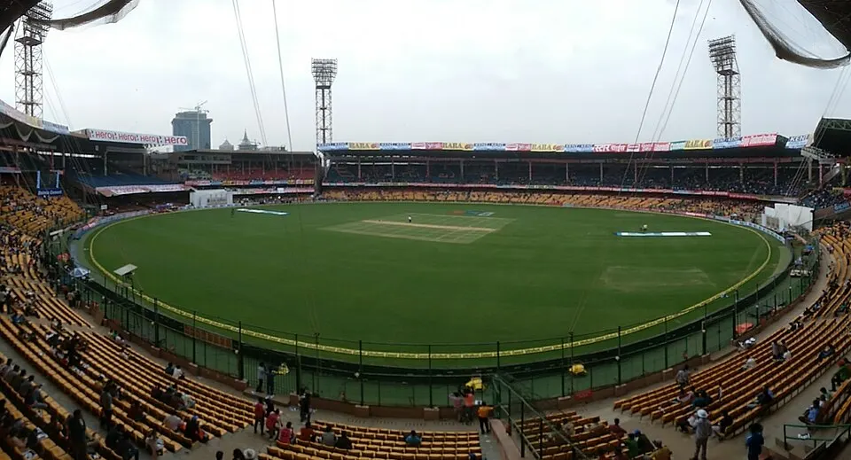 A wide view of a cricket ground with yellow seats, floodlight towers and a green outfield.