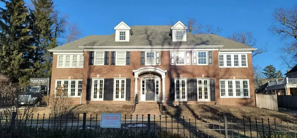 A large red-brick house with white trim, black shutters and a pillared front door, seen from across an iron fence.