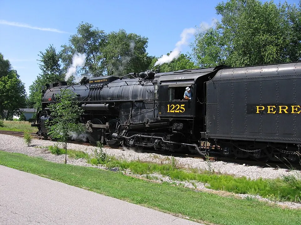 A black steam locomotive with yellow numbers on the cab pulling a tender, with white steam rising among green trees.