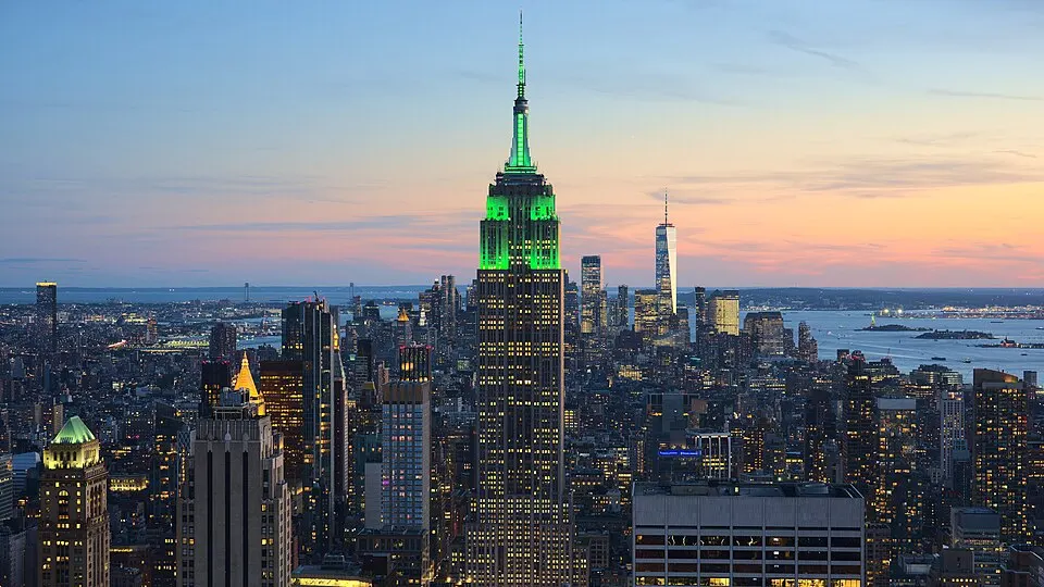 A tall skyscraper with a lit green spire rising above a dense city skyline at dusk, with water in the distance.
