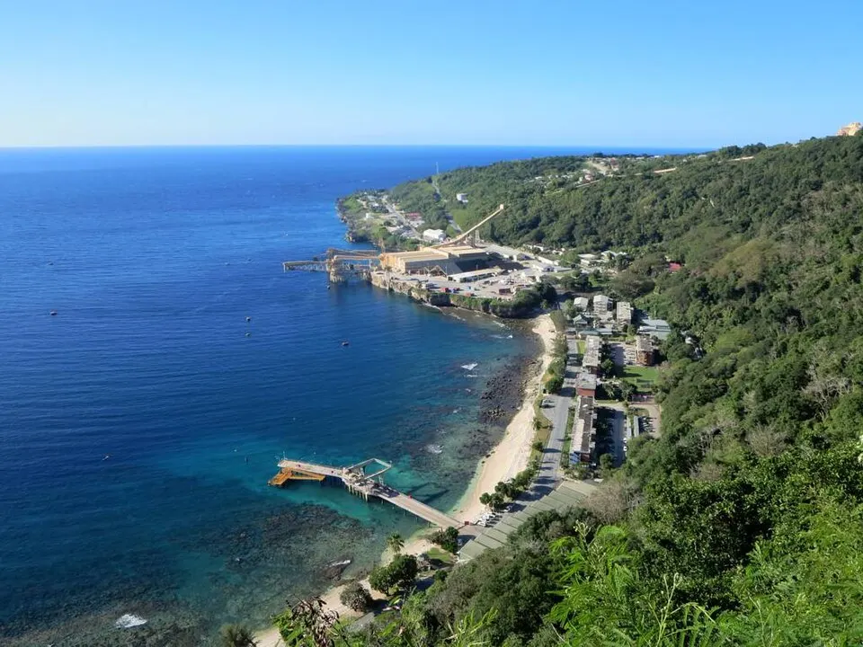 A steep green hillside above a curving bay of deep blue water, with a pier, a small beach and a few buildings along the shore.