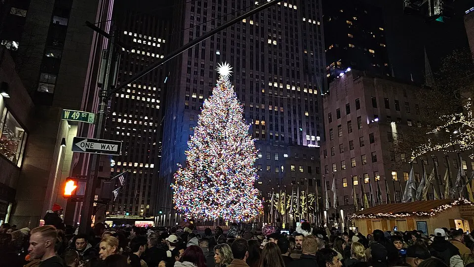 A huge Christmas tree covered in colourful lights and topped with a star glows between skyscrapers above a crowd.