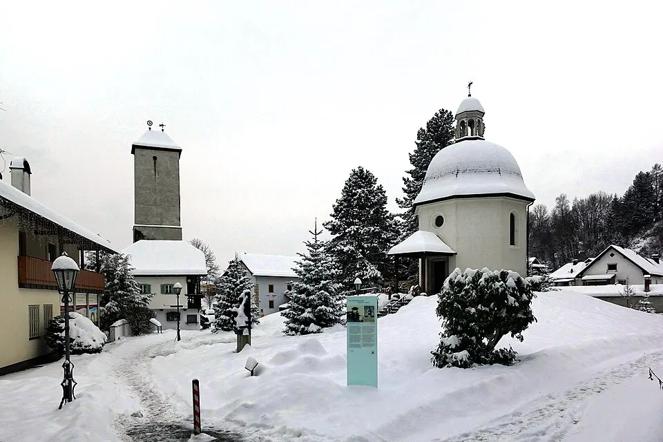 A small round white chapel with a little bell tower stands in a snowy village with fir trees.