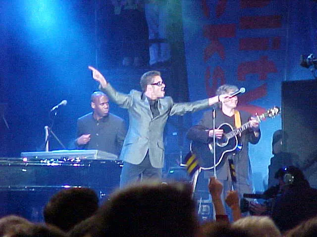 A man in glasses and a grey suit points one arm out while singing on a blue-lit stage, with a keyboard player and guitarist beside him.