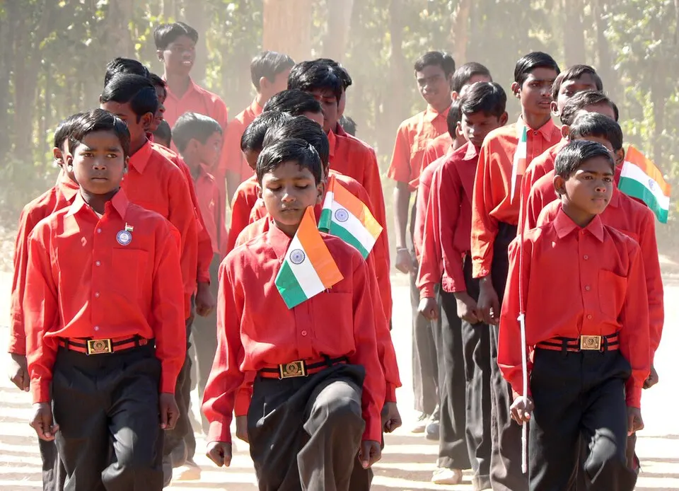 लाल कमीज़ पहने बच्चे हाथों में तिरंगे झंडे लिए कतार में चल रहे हैं।