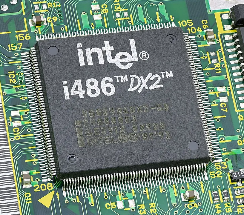 A square black computer chip with rows of tiny metal pins mounted on a green circuit board.