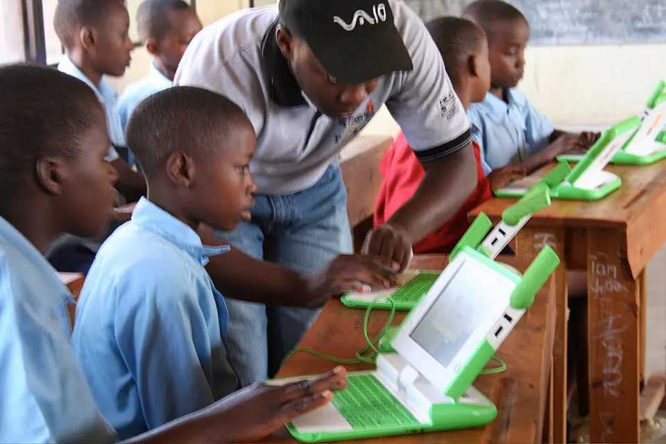 Schoolchildren in blue uniforms using small green-and-white laptops at wooden desks while a man helps one of them.