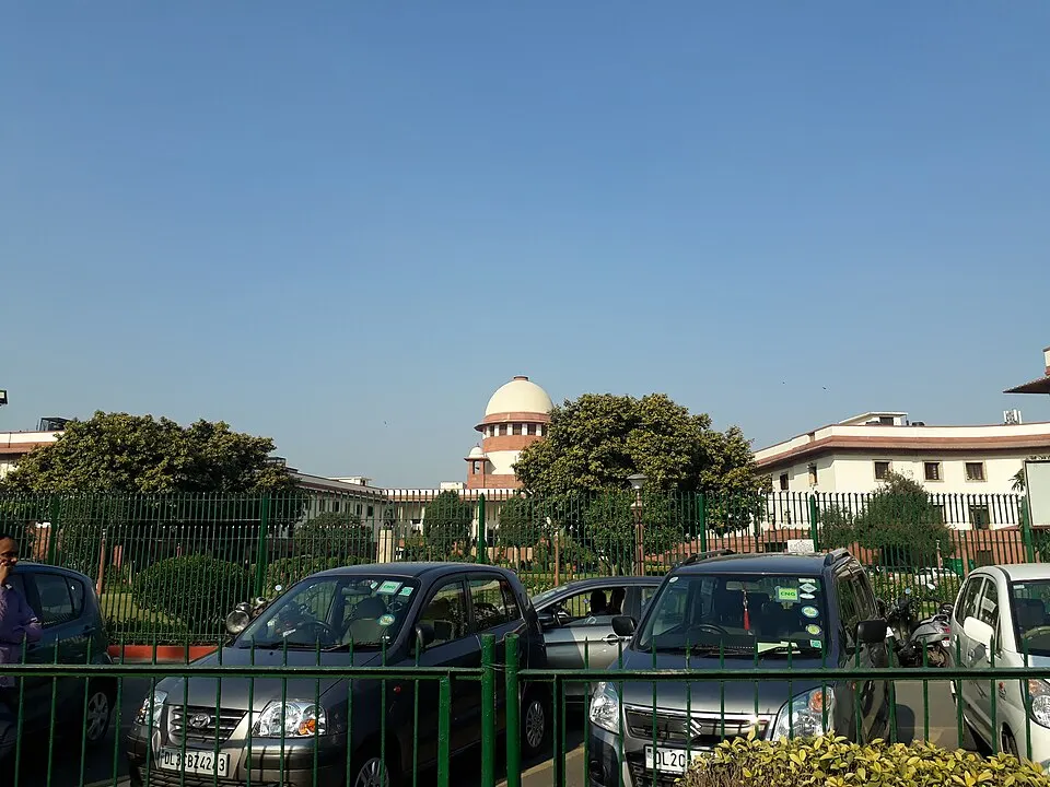 A white building with a pink-banded domed tower behind a green iron fence and parked cars.