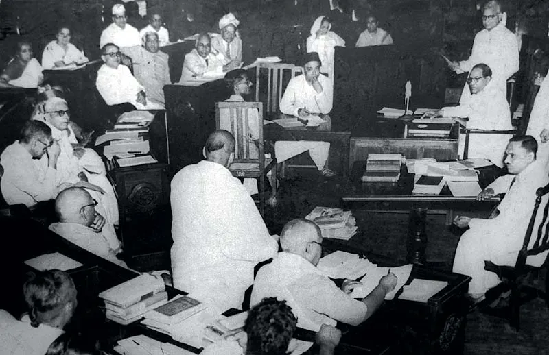 A black and white photo of men in white clothes seated at desks piled with books in a legislative chamber.