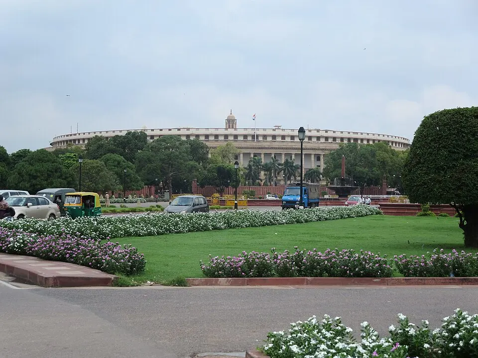 A large circular colonnaded building across a green lawn with flowering shrubs, autorickshaws and cars on the road.