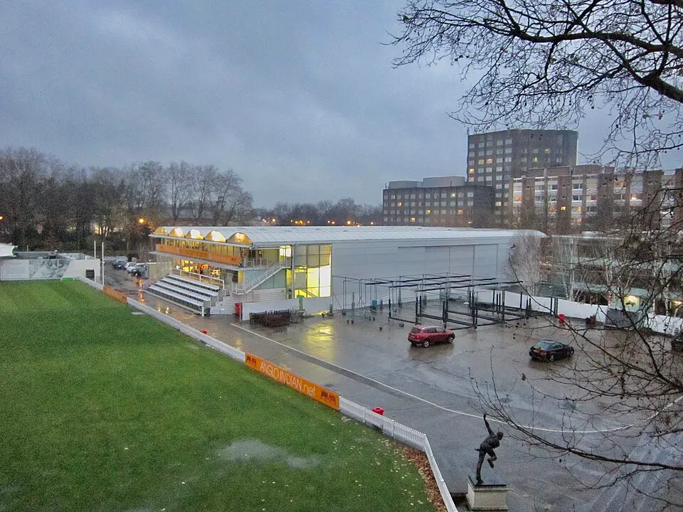 A view from a glass-fronted box over a green cricket ground with a red-brick pavilion on the far side.