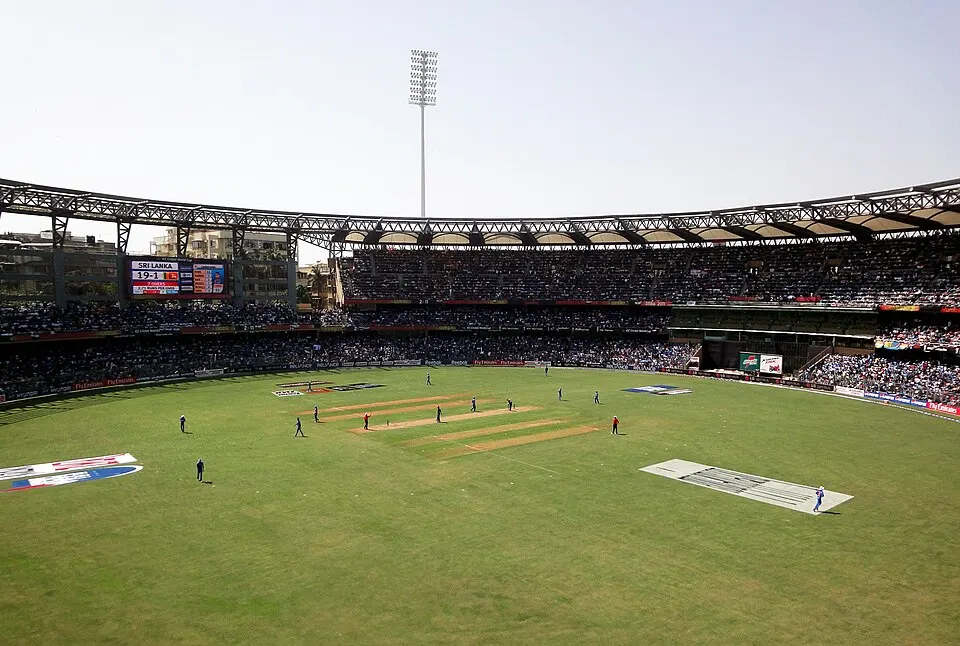 A packed cricket stadium under a bright sky with players on the field and a scoreboard at one end.