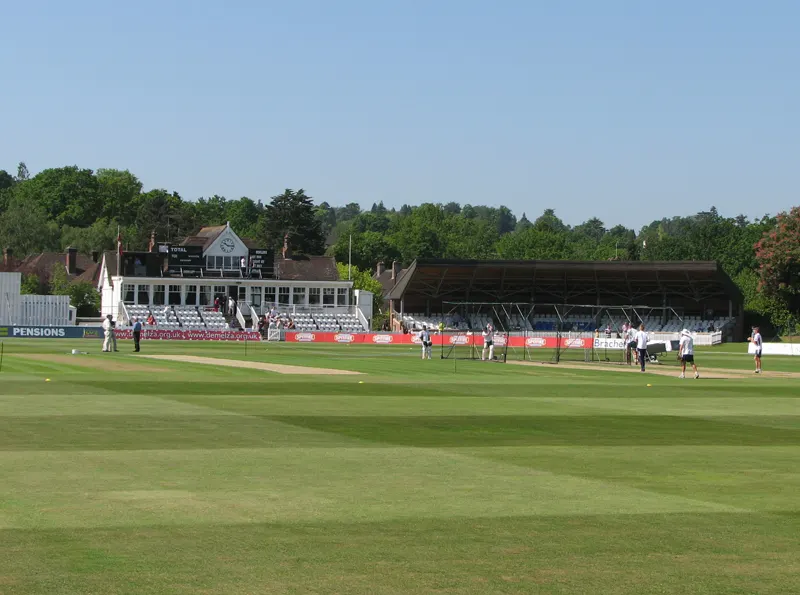 A small English cricket ground with a white pavilion with a clock, a covered stand and players on the grass.