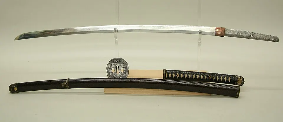 A long, slightly curved Japanese sword with its metal guard and dark lacquered scabbard laid out in a museum.